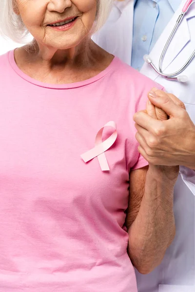 Vista ritagliata del medico e della donna anziana con nastro rosa sulla t-shirt che tiene le mani isolate sul bianco — Foto stock