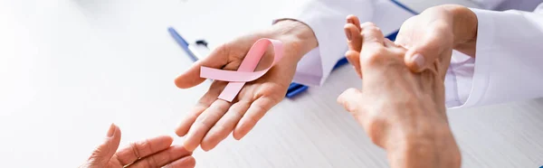 Panoramic shot of doctor holding patient hands and showing pink ribbon — Stock Photo