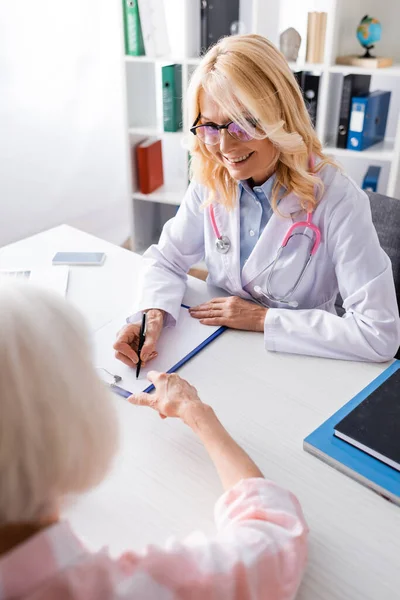 Médecin en manteau blanc écrit sur presse-papiers assis près du patient à la clinique — Photo de stock