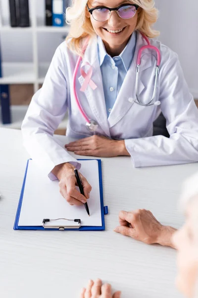 Enfoque selectivo del médico escribiendo en el portapapeles y mirando al paciente en la clínica - foto de stock