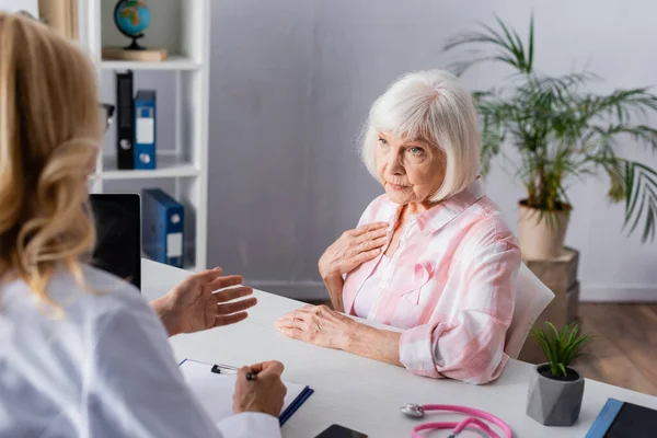 Focus selettivo della donna anziana con nastro rosa seduta vicino al medico in clinica — Foto stock