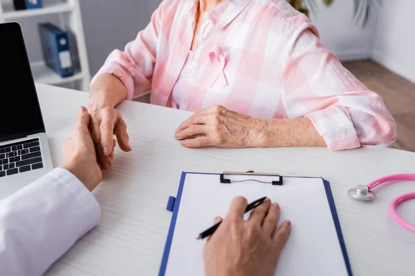 Vista recortada del médico sosteniendo la mano del paciente cerca de la computadora portátil y portapapeles - foto de stock