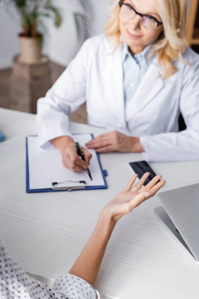 Selective focus of patient sitting near doctor writing on clipboard at workplace — Stock Photo