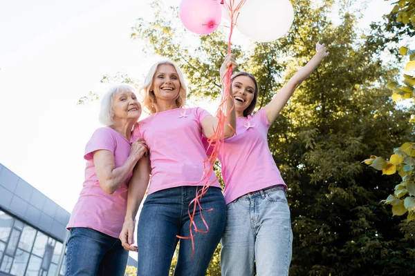 Vue en angle bas de trois femmes en t-shirts avec rubans roses tenant des ballons — Photo de stock