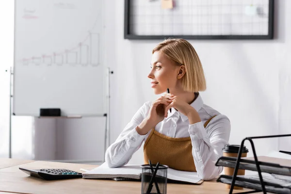 Femme d'affaires blonde regardant loin tout en étant assis au bureau avec papeterie dans le bureau sur fond flou — Photo de stock