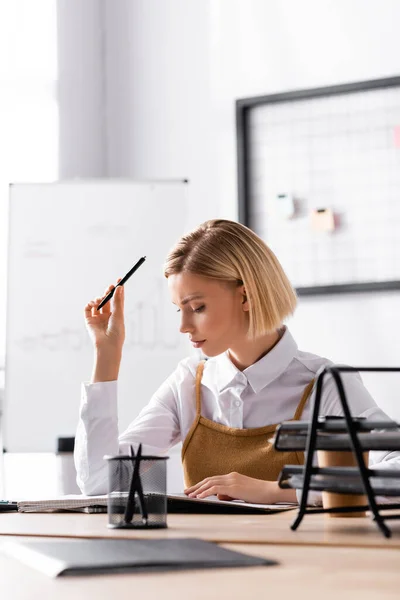 Konzentrierte blonde Frau mit Stift blickt auf Notizblock, während sie am Schreibtisch im Büro auf verschwommenem Hintergrund sitzt — Stockfoto