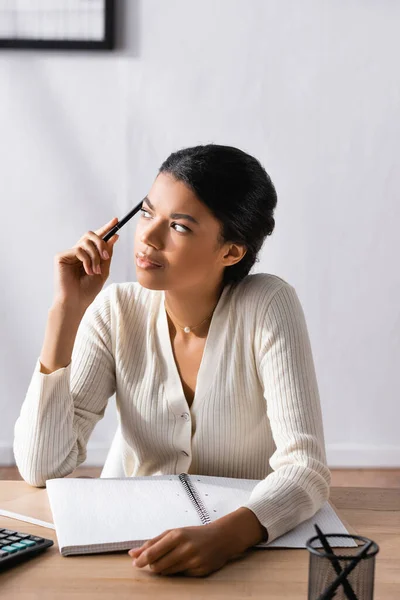 Pensativa mujer afroamericana sosteniendo la pluma y mirando hacia otro lado mientras se sienta en el escritorio en la oficina sobre un fondo borroso - foto de stock