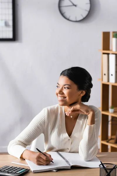 Mujer afroamericana feliz con bolígrafo, mirando hacia otro lado mientras está sentado en el escritorio con papelería en la oficina sobre fondo borroso - foto de stock