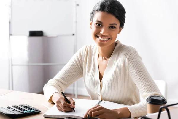 Glückliche afrikanisch-amerikanische Frau mit Stift, die in die Kamera schaut, während sie auf leerem Notizbuch im Büro auf verschwommenem Hintergrund schreibt — Stockfoto