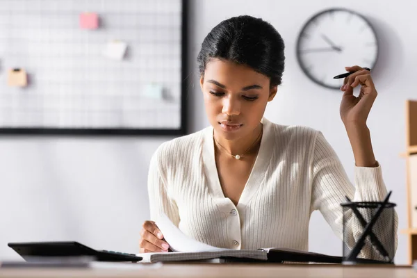 Oberflächenebene einer fokussierten Afroamerikanerin, die auf ein Notizbuch blickt, während sie am Schreibtisch im Büro auf verschwommenem Hintergrund sitzt — Stockfoto