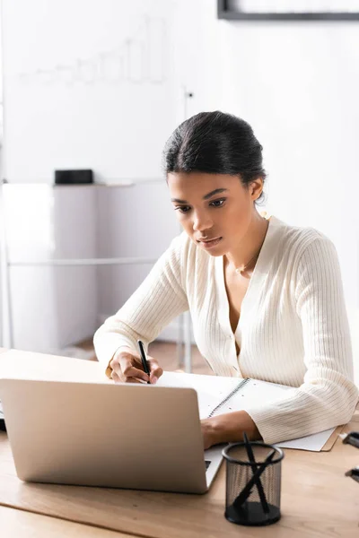 Focused african american woman looking at laptop while writing on blank notebook with blurred flipchart on background — Stock Photo