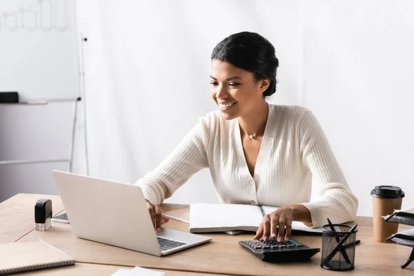 Heureuse femme afro-américaine regardant ordinateur portable tout en comptabilisant sur la calculatrice au bureau sur fond flou — Photo de stock