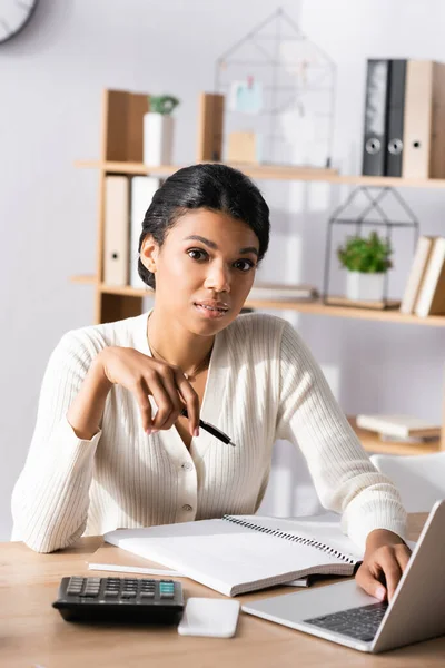 Mujer de negocios afroamericana con pluma mirando a la cámara mientras escribe en el ordenador portátil en un fondo borroso - foto de stock