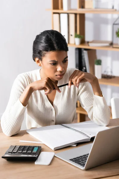 Задумчивая африканская американка смотрит на ноутбук, сидя за столом в офисе на размытом фоне — стоковое фото