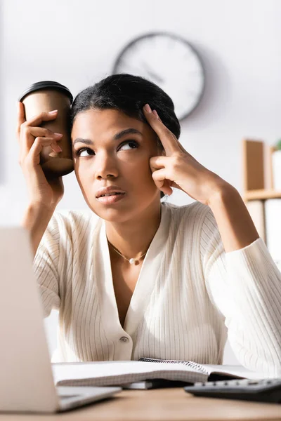 Tired african american woman looking away and holding cup of coffee near head in office on blurred foreground — Stock Photo