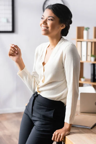 Feliz mujer de negocios afroamericana mirando hacia otro lado mientras se apoya en la mesa en la oficina sobre un fondo borroso - foto de stock