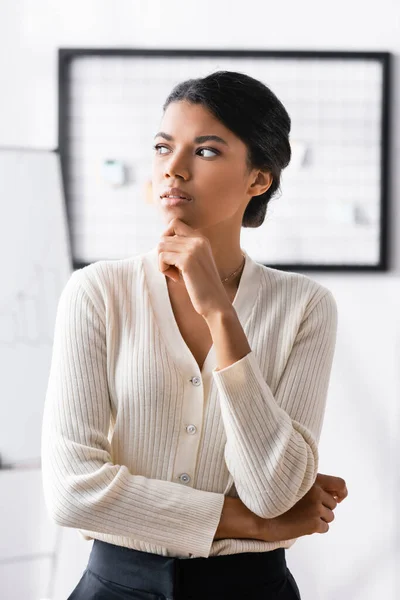 Nachdenkliche Afroamerikanerin mit einer Hand am Kinn, die wegschaut, während sie im Büro auf verschwommenem Hintergrund steht — Stockfoto