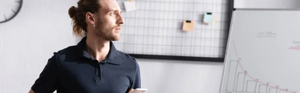 Fiducioso uomo d'affari distogliendo lo sguardo mentre in piedi vicino a flip chart e organizzatore di mesh in ufficio, banner — Foto stock