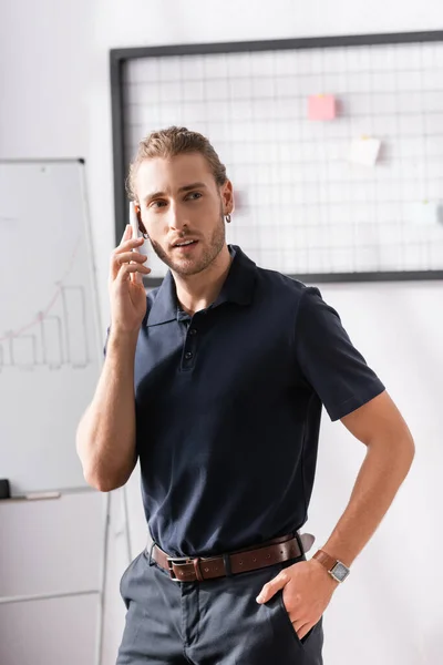 Hombre de negocios seguro con la mano en el bolsillo mirando hacia otro lado mientras habla por teléfono en la oficina sobre fondo borroso - foto de stock