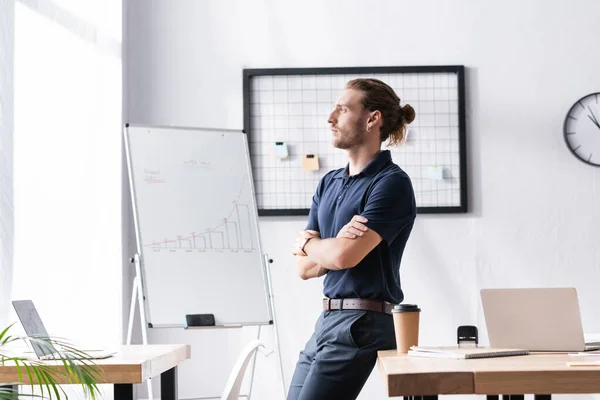 Selbstbewusster Geschäftsmann mit verschränkten Armen, der wegschaut, während er sich im Büro auf den Tisch lehnt — Stockfoto