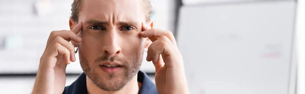 Exhausted young adult man looking at camera and touching head with hands on blurred background, banner — Stock Photo