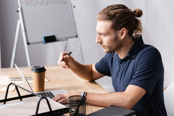Trabajador de oficina enfocado con bolígrafo mirando el portátil mientras está sentado en la mesa del lugar de trabajo sobre un fondo borroso - foto de stock