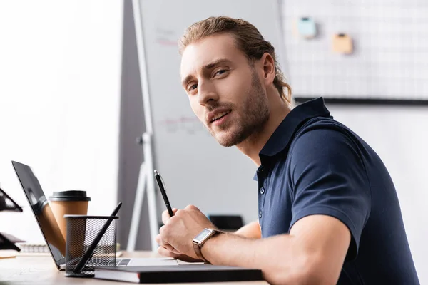Confident office worker looking at camera while sitting near laptop at workplace table on blurred background — Stock Photo