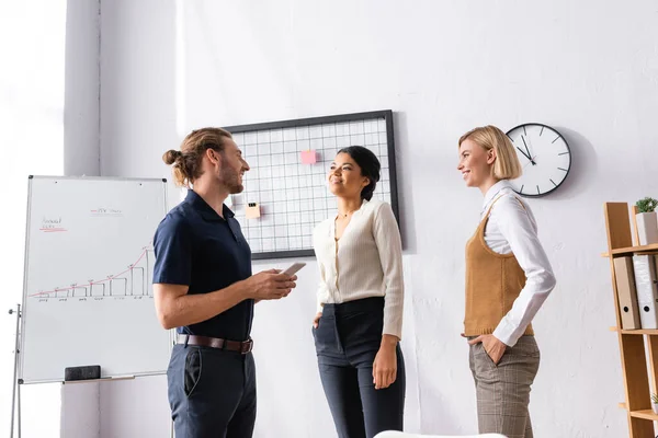 Smiling multicultural office workers looking at each other while standing near mesh organizer and flipchart at workplace — Stock Photo