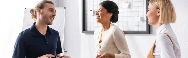 Happy multicultural office workers talking while standing near mesh organizer and flipchart at workplace, banner — Stock Photo