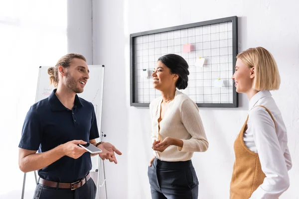 Cheerful multiethnic office workers talking while standing near mesh organizer and flipchart at workplace — Stock Photo