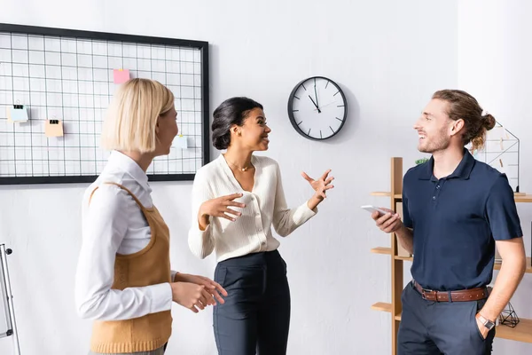 Heureux travailleurs de bureau multiethniques gesticuler et parler tout en se tenant près de l'organisateur de maille avec des autocollants vierges sur le lieu de travail — Photo de stock