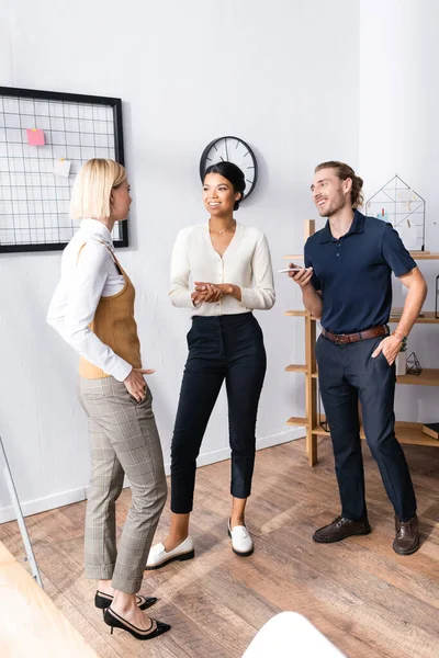 Multiethnic smiling office workers talking while standing near mesh organizer with blank stickers at workplace — Stock Photo
