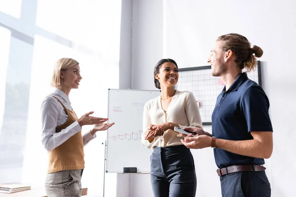 Fröhliche multikulturelle Büroangestellte gestikulieren und schauen einander an, während sie am Arbeitsplatz neben Flipchart mit Grafik stehen — Stockfoto