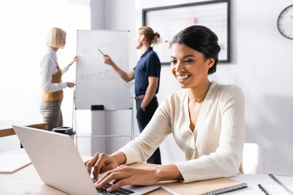 Lächelnde Afroamerikanerin tippt am Laptop, während sie am Arbeitsplatz mit verschwommenen Kollegen im Hintergrund sitzt — Stockfoto