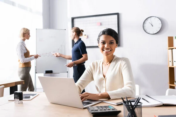 Femme afro-américaine souriante regardant la caméra, tout en tapant sur un ordinateur portable avec des employés de bureau flous sur fond — Photo de stock