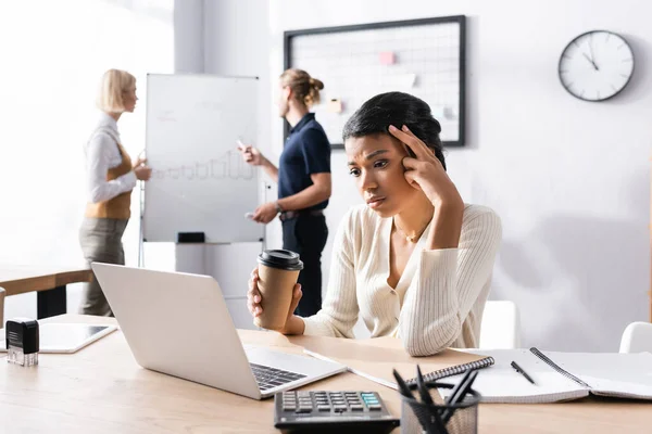 Donna afroamericana stanca con tazza di carta che guarda il computer portatile sul posto di lavoro con impiegati offuscati sullo sfondo — Foto stock