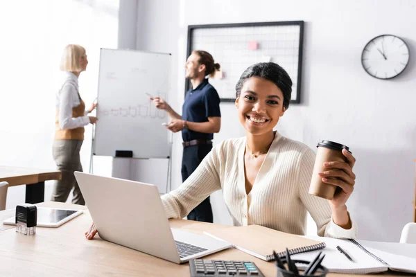 Happy african american woman with paper cup looking at camera while sitting at table on blurred background — Stock Photo