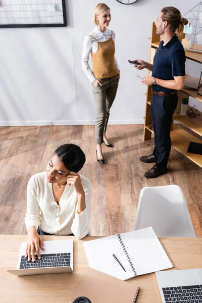 Overhead-Ansicht einer afrikanisch-amerikanischen Frau, die mit einem Notizbuch am Schreibtisch sitzt, während Büroangestellte im Hintergrund sprechen — Stockfoto