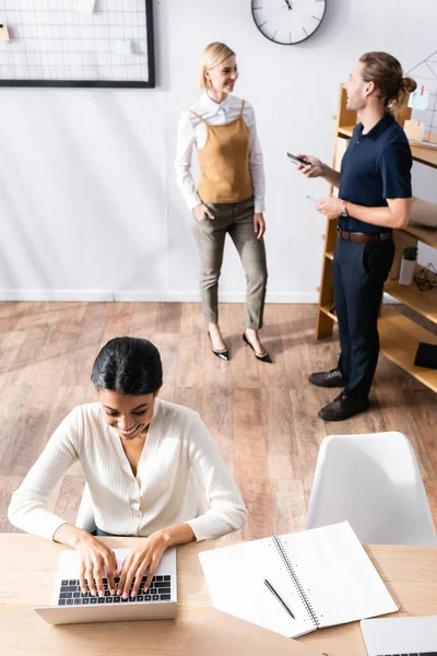 Visão aérea da mulher americana africana feliz digitando no laptop enquanto está sentado na mesa com trabalhadores de escritório falando em segundo plano — Fotografia de Stock