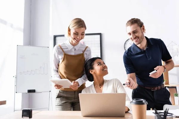 Happy office workers standing near african american woman sitting at table with laptop at workplace — Stock Photo