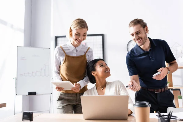 Heureux employés de bureau debout près de femme afro-américaine assis à la table avec ordinateur portable sur le lieu de travail — Photo de stock