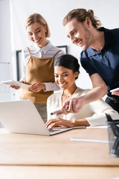 Man looking at laptop and pointing with marker, while standing near multicultural female office workers at workplace — Stock Photo