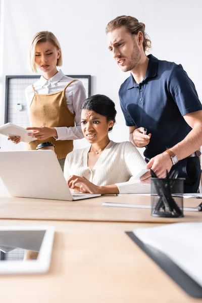 Trabajadores de oficina escépticos de pie cerca de la mujer afroamericana y mirando a la computadora portátil en la mesa con papelería borrosa en primer plano - foto de stock