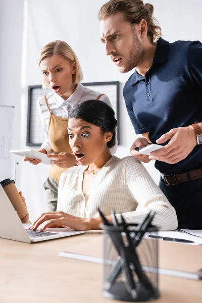 Impresionados trabajadores de oficinas multiculturales mirando a la computadora portátil en el lugar de trabajo con plumas borrosas titular en primer plano - foto de stock