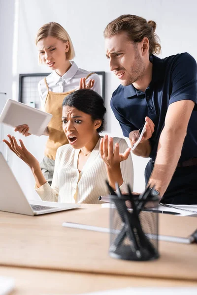 Lavoratori d'ufficio multiculturali offesi che gesticolano mentre guardano laptop sul posto di lavoro con portapenne sfocato in primo piano — Foto stock