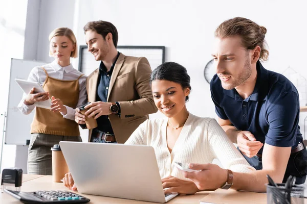 Trabalhadores de escritório multiculturais sorridentes olhando para laptop na mesa, enquanto colegas trabalhando em segundo plano no escritório — Fotografia de Stock