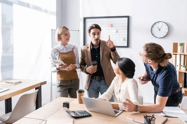 Multicultural office workers looking at excited colleague with idea gesture while standing in office near workplaces — Stock Photo