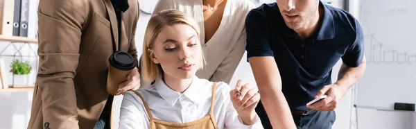 Focused woman pointing with pen with multicultural office workers standing behind on blurred background, banner — Stock Photo