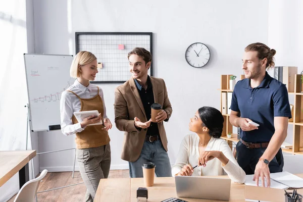 Skeptical multicultural office workers looking at smiling colleagues speaking and gesturing while standing in office near workplaces — Stock Photo
