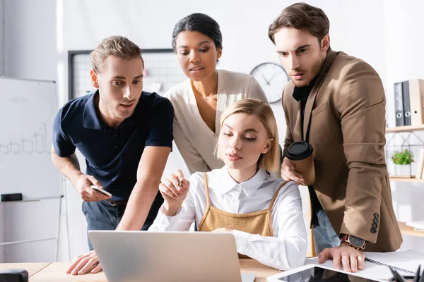 Konzentrierte Frau zeigt mit Stift, während sie am Tisch sitzt, hinter dem fokussierte multikulturelle Büroangestellte auf verschwommenem Hintergrund stehen — Stockfoto